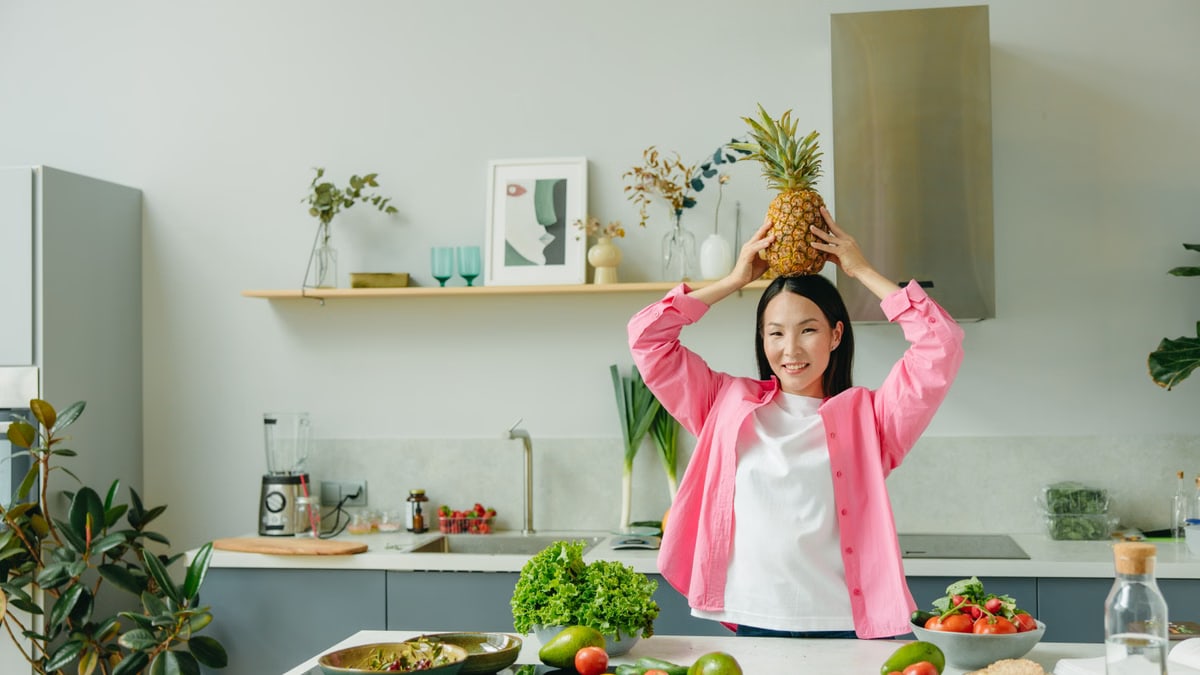 Asian woman in pink overshirt holding a pineapple in a modern kitchen with fresh produce and cheerful ambiance.